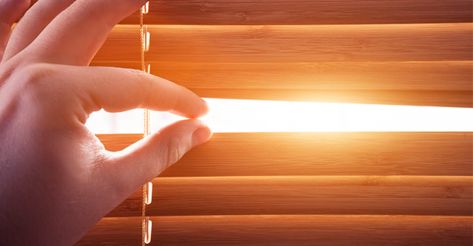 Hand opening wooden window blinds to let in bright sunlight